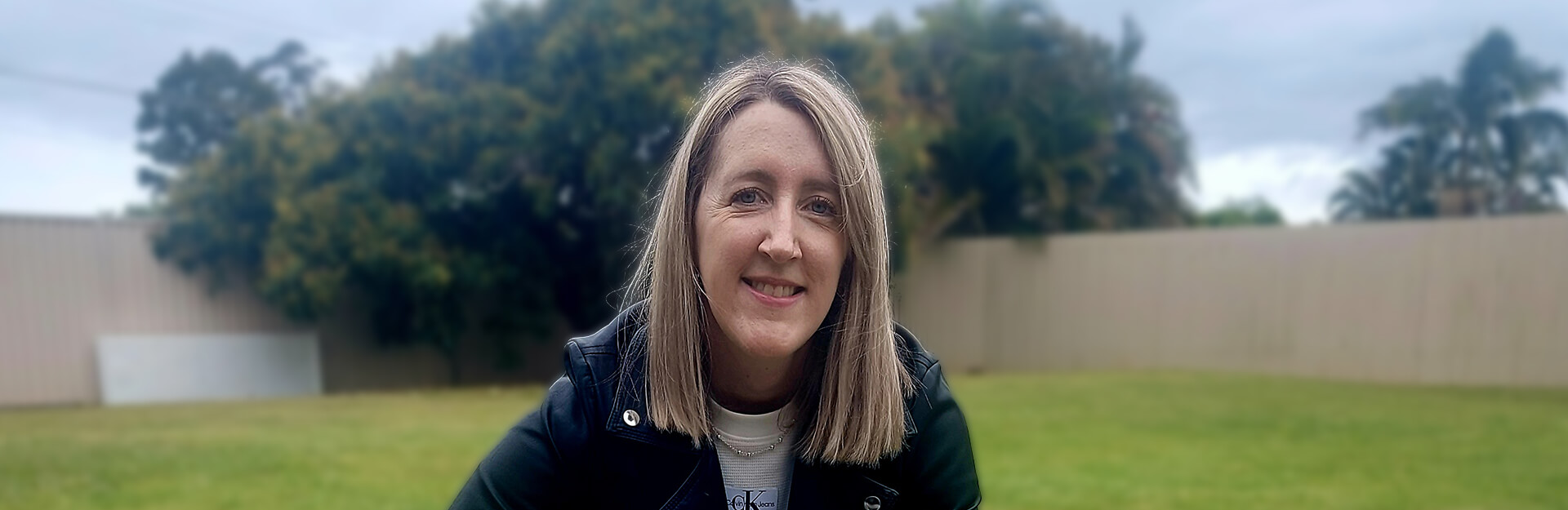 A woman with long hair smiles while sitting outdoors in a green yard, with trees and a fence in the background.