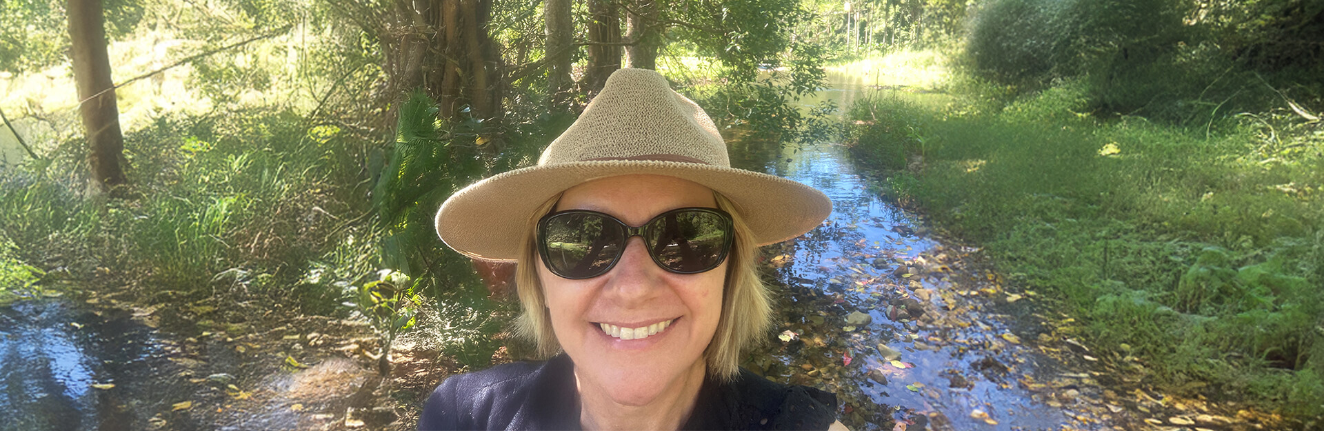 A person wearing sunglasses and a wide-brimmed hat smiles in a lush, green outdoor setting near a stream.