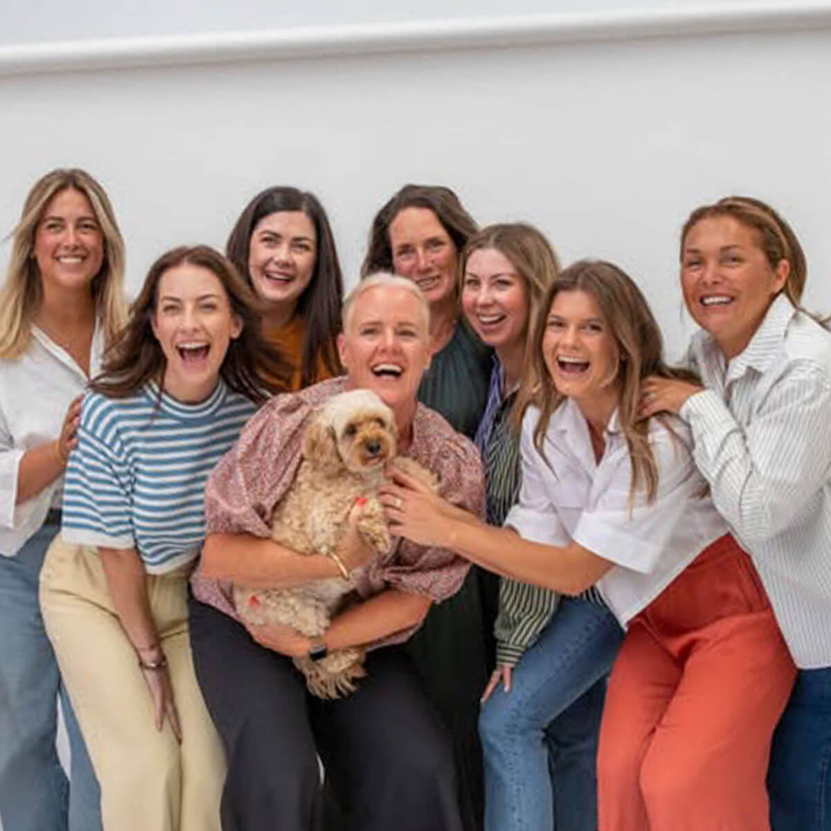 A group of nine women smiling and posing together, one holding a small dog. Bright, friendly atmosphere.