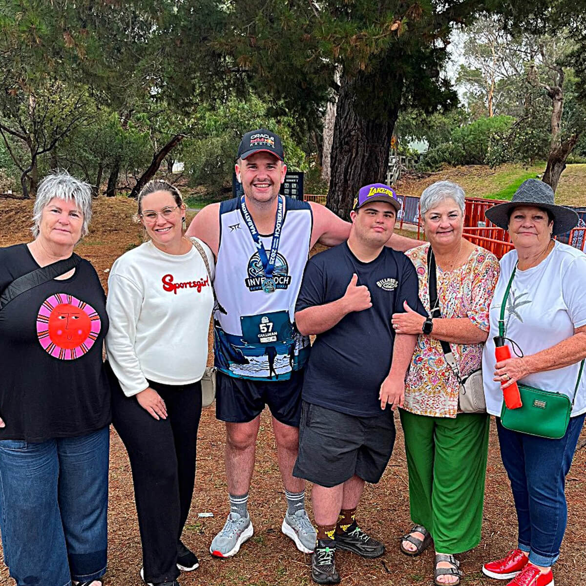 A group of six people, smiling and posing together outdoors, with trees in the background. One person is wearing a medal.