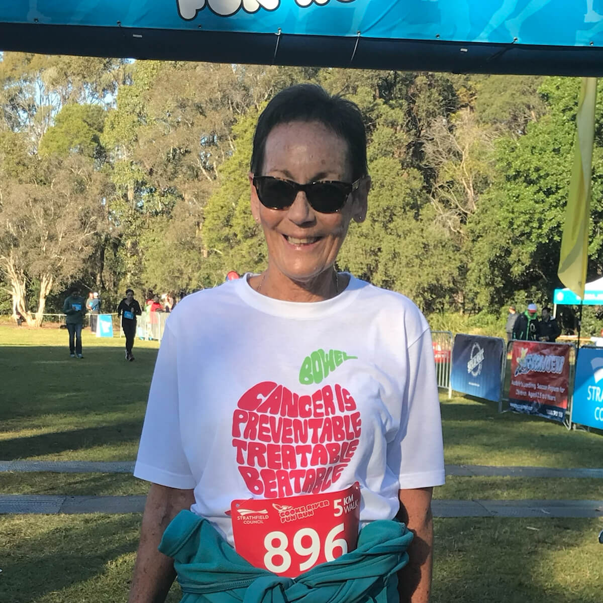 A smiling woman in sunglasses stands in front of a race banner, wearing a shirt with a cancer awareness message and a race bib.