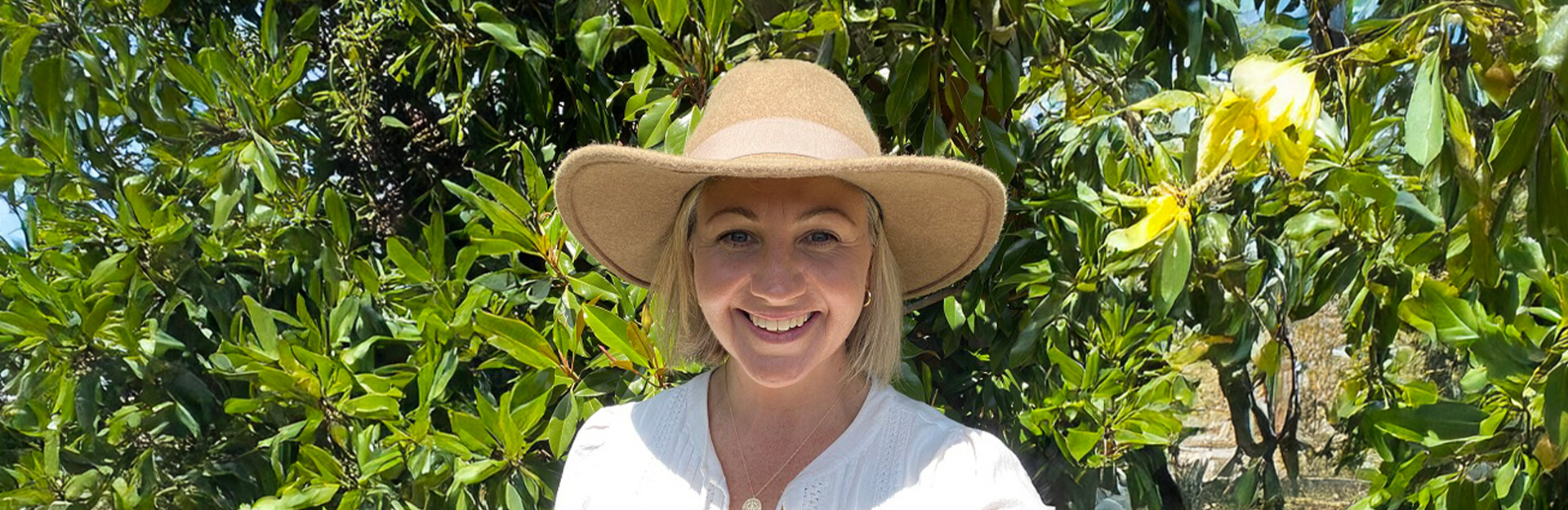 A smiling woman wearing a wide-brimmed hat stands in front of lush green foliage.