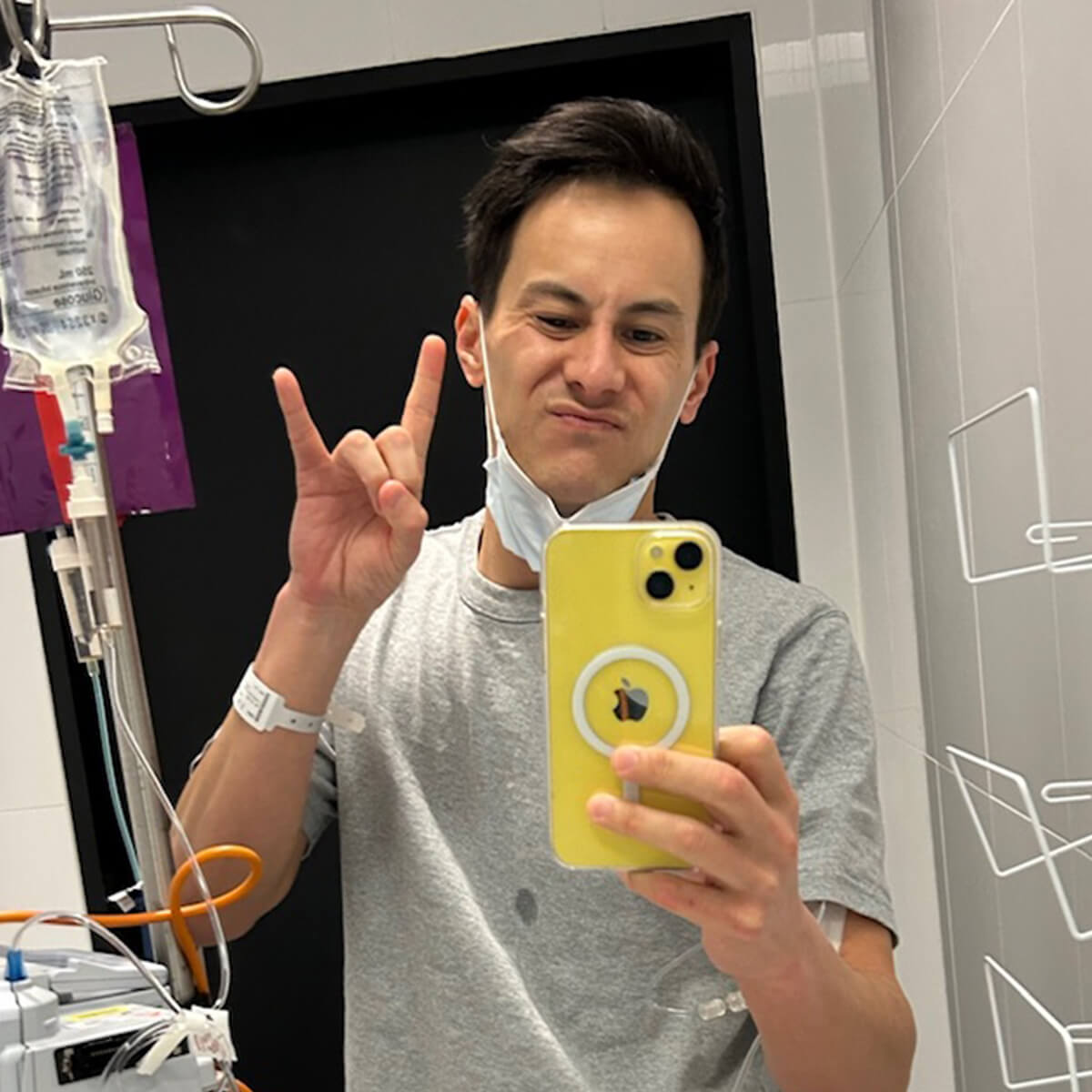 A person in a hospital setting takes a selfie, wearing a mask and showing a hand gesture, with an IV pole nearby.