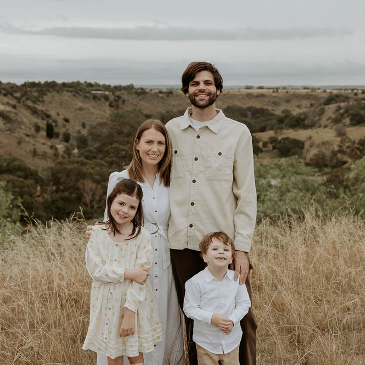 A family of four stands together in a grassy field with hills in the background, smiling at the camera.
