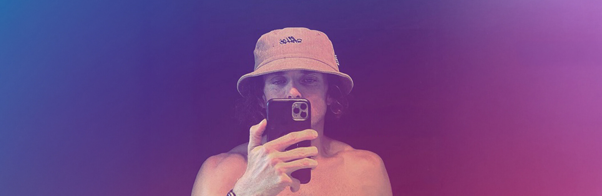 A person with curly hair wearing a bucket hat takes a selfie, showing their bare shoulders and holding a smartphone.
