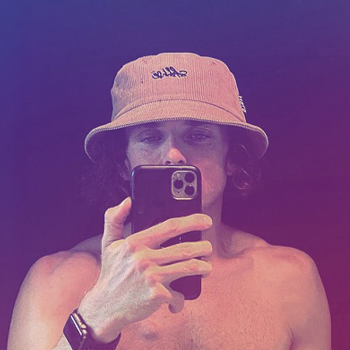 A person with curly hair wearing a bucket hat takes a selfie, showing their bare shoulders and holding a smartphone.