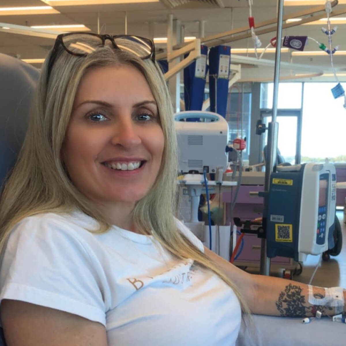 A smiling woman with long blonde hair sits in a medical setting, wearing a white shirt and connected to equipment.