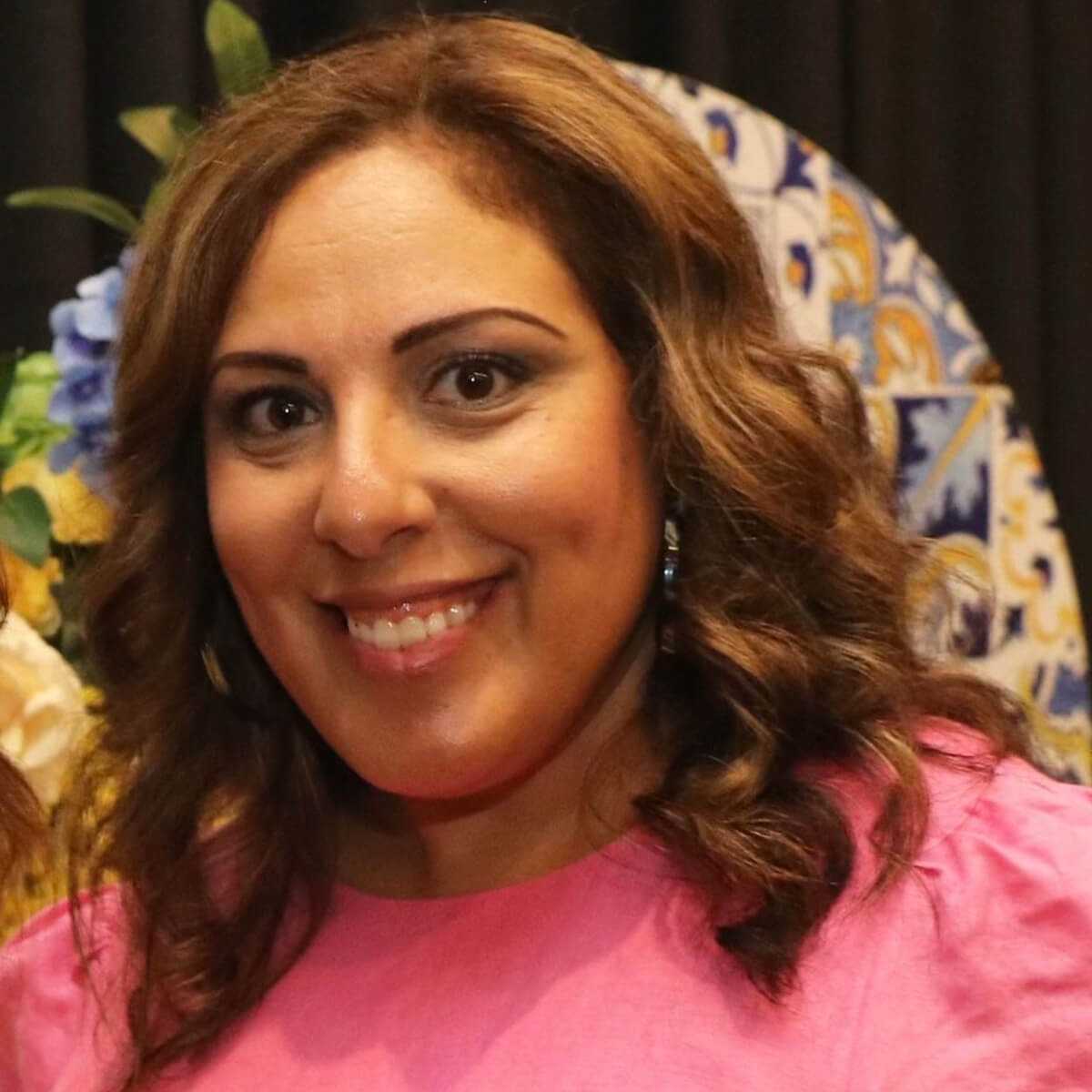 A woman with curly brown hair smiles, wearing a pink top, with a decorative background of flowers and patterns.