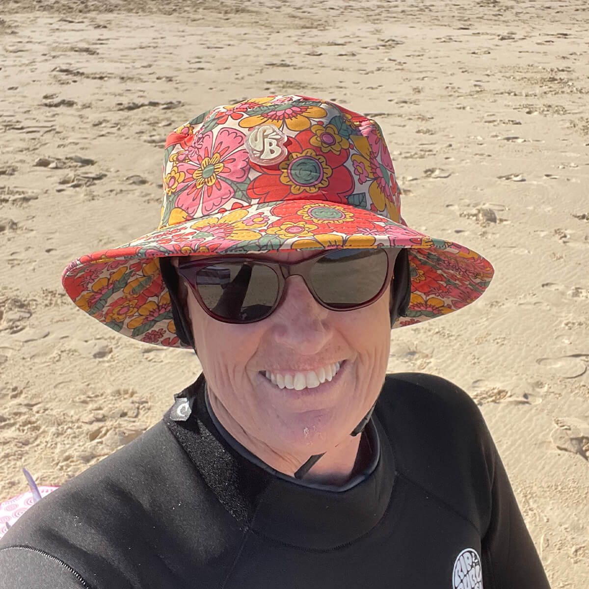 A person wearing a floral bucket hat and sunglasses smiles at the beach, with sand in the background.