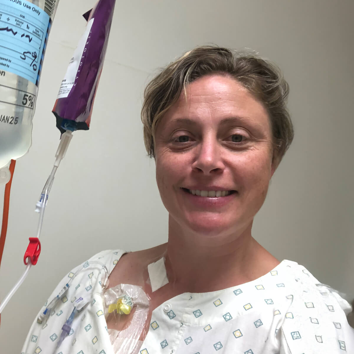 A smiling woman in a hospital gown with an IV drip attached to her arm.
