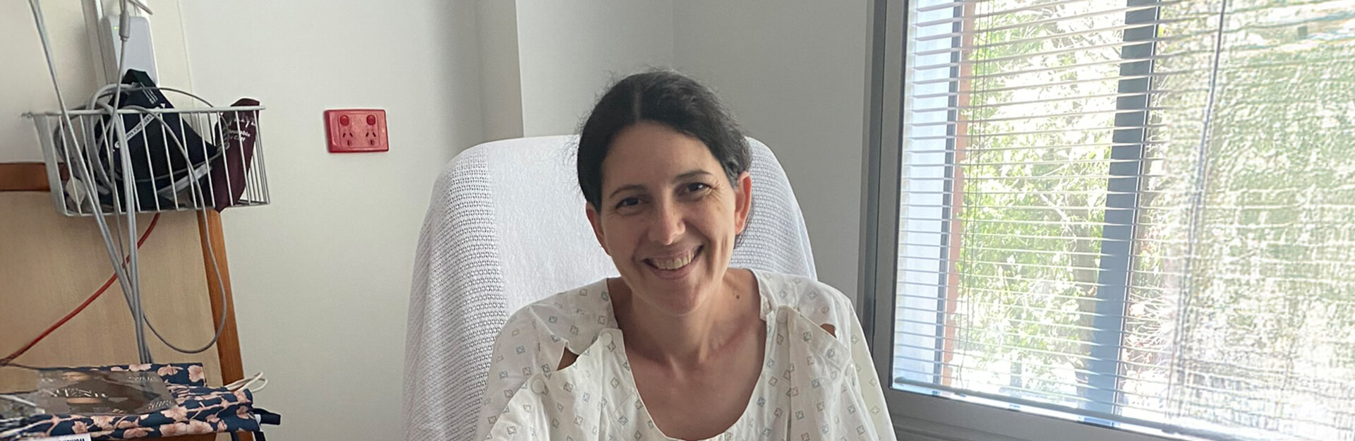 A woman in a hospital gown smiles while sitting on a bed next to a window with blinds.