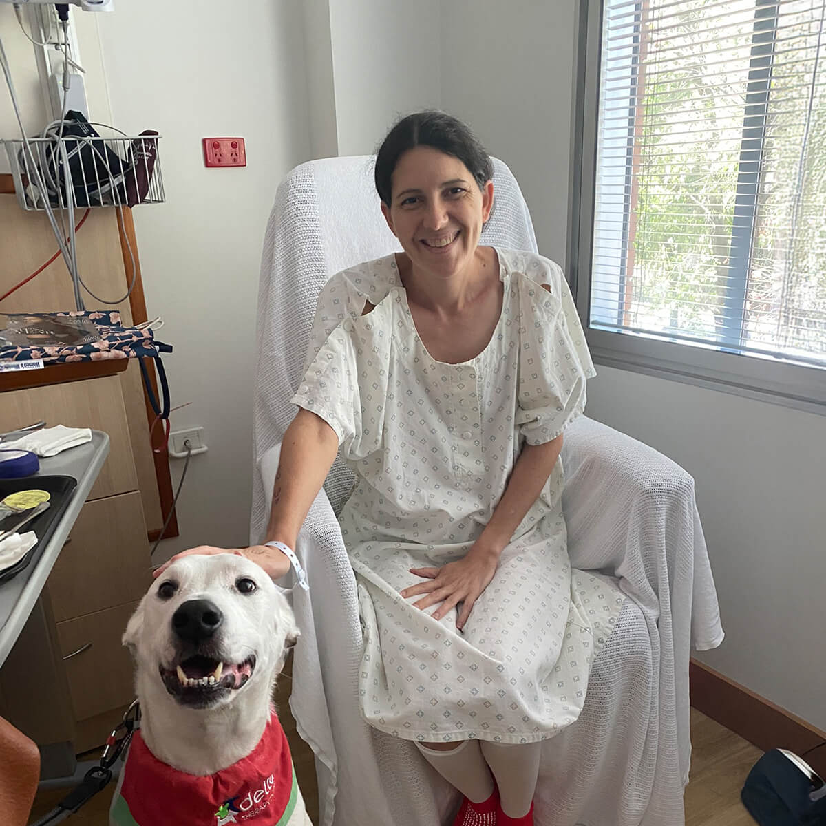 A woman in a hospital gown sits in a chair, smiling at a dog beside her. There are medical supplies on a nearby table.