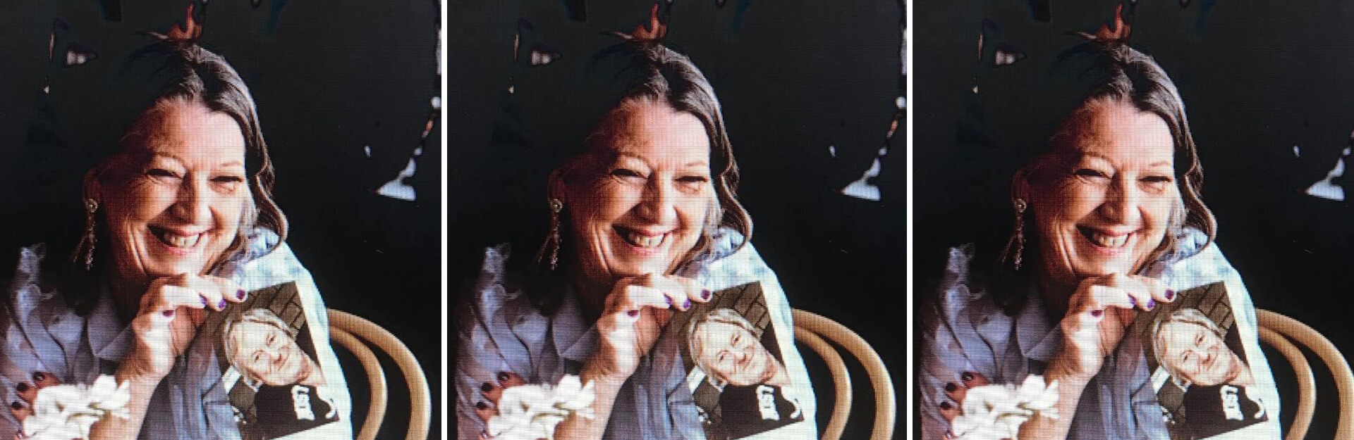A smiling woman holds a photo while sitting at a table, with a flower arrangement nearby.