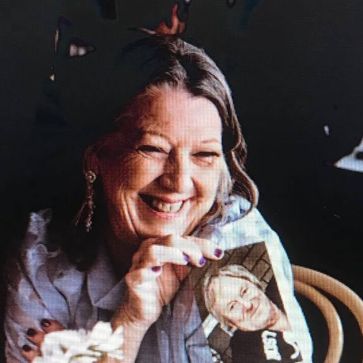 A smiling woman holds a photo of another person, sitting at a table with flowers.