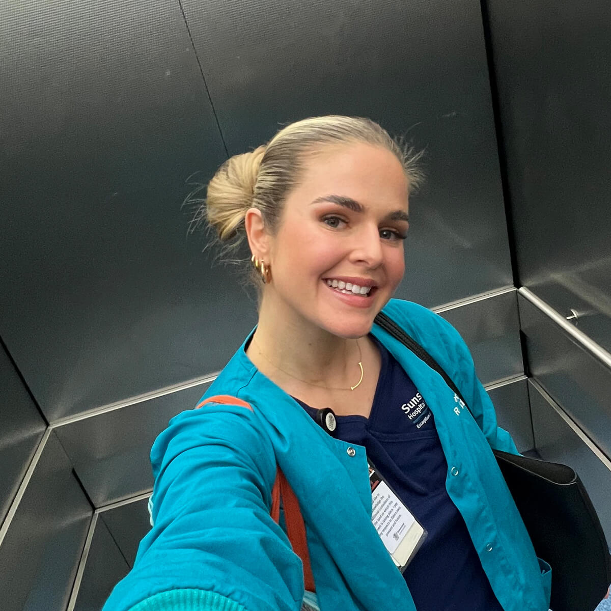 A smiling woman in a teal jacket and dark shirt poses for a selfie in an elevator.