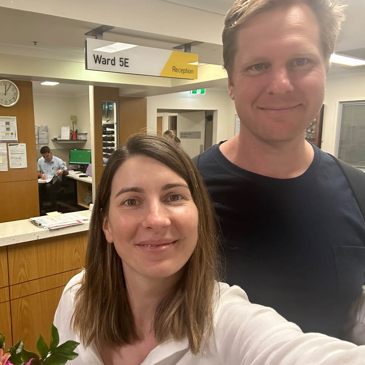 A woman and a man smile for a selfie in a hospital reception area with a sign for "Ward 5E" visible.