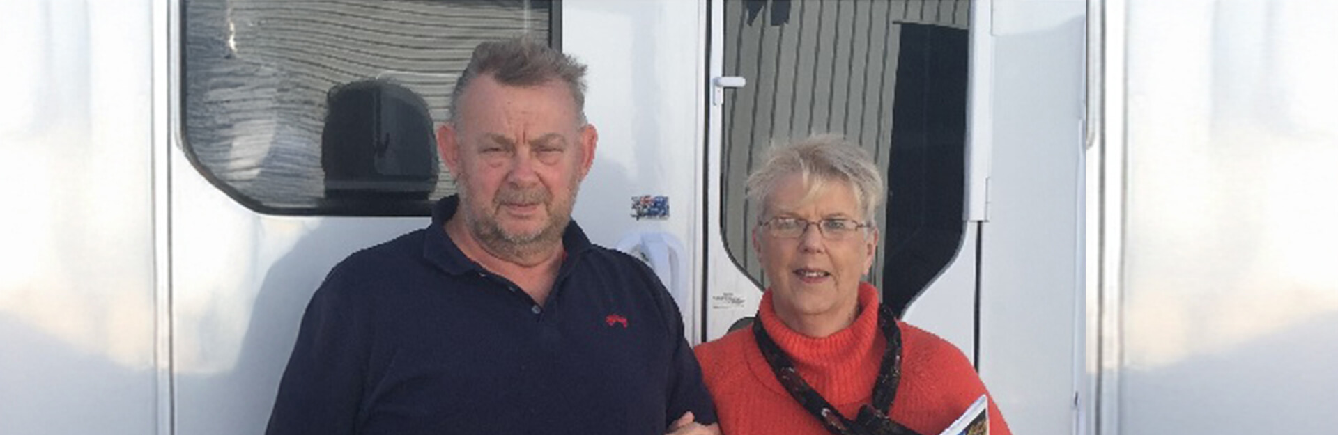 A man and woman stand together outside a trailer, both smiling and dressed casually.