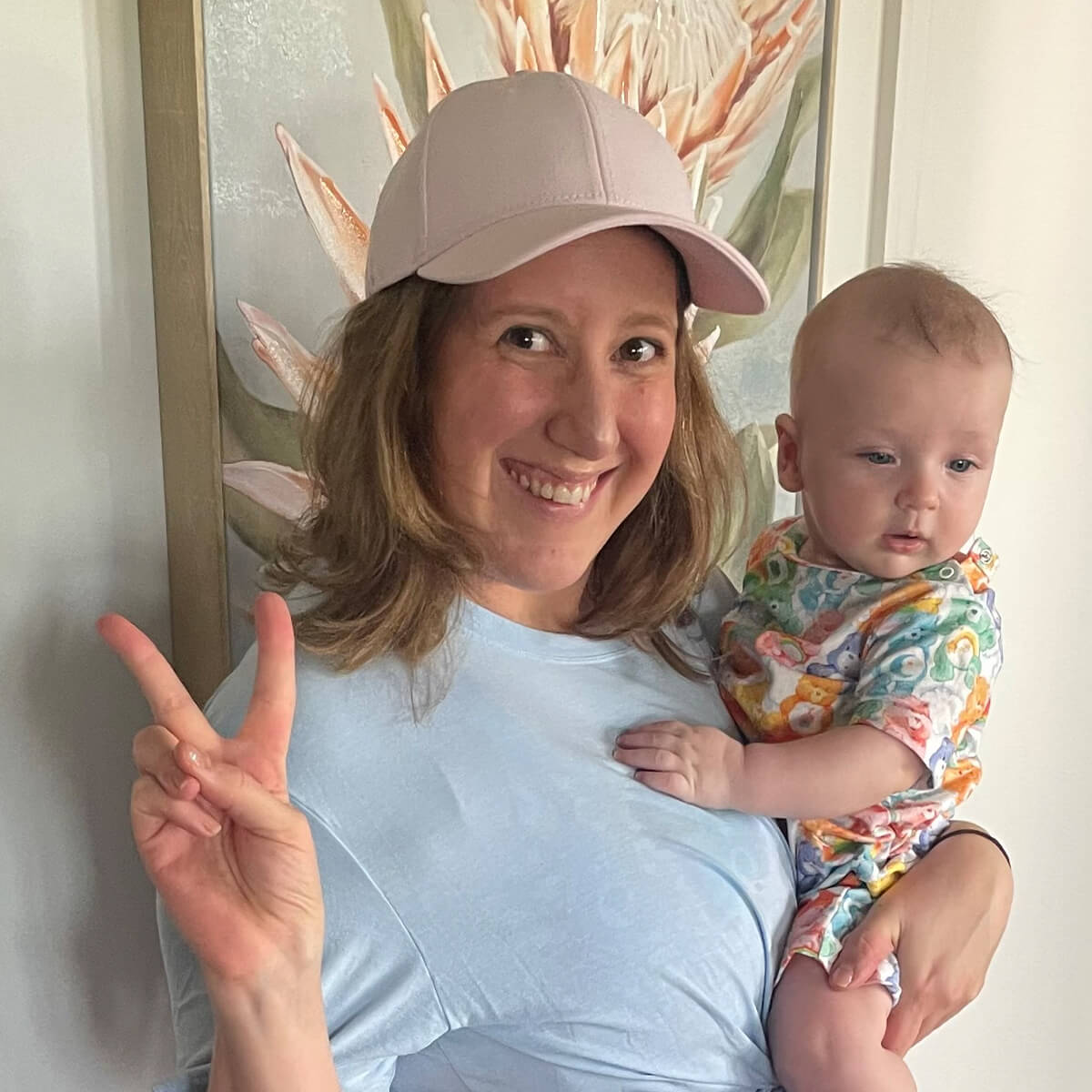 A smiling woman wearing a pink cap holds a baby in colorful attire, with a floral painting in the background.