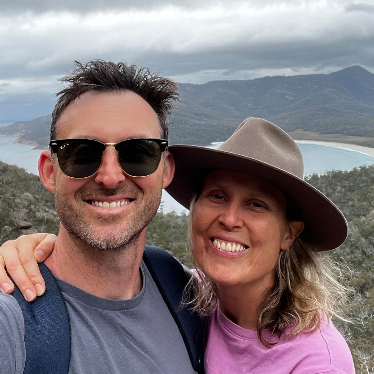 A man and a woman smile for a selfie with a scenic mountainous landscape and ocean in the background.