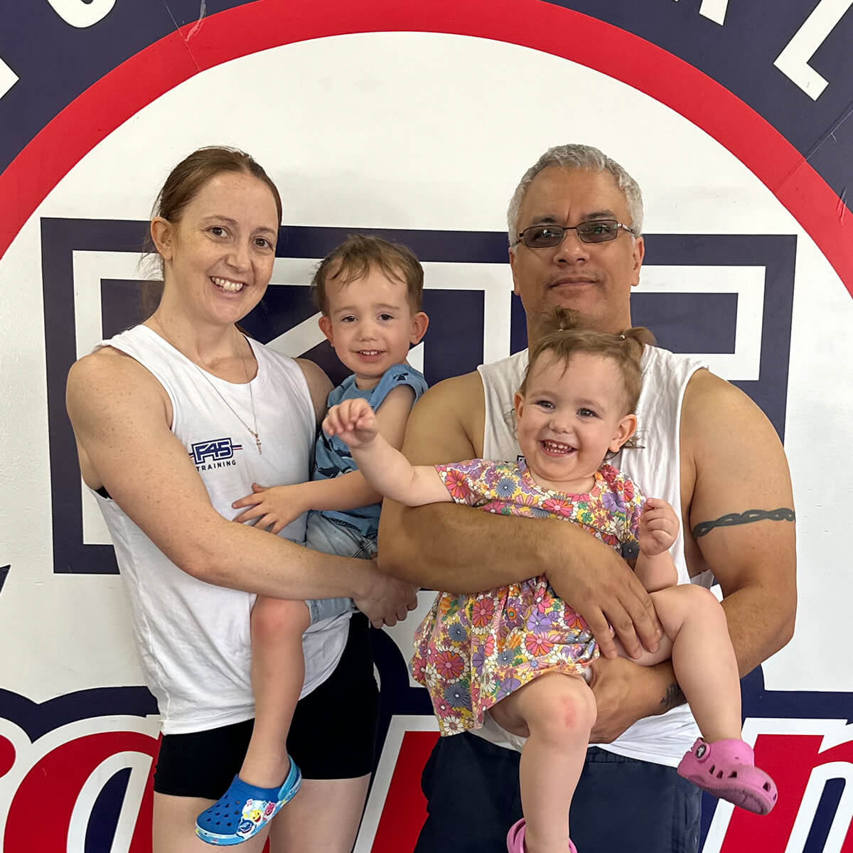 A smiling family of four poses together in front of a colorful wall, with two young children being held by their parents.