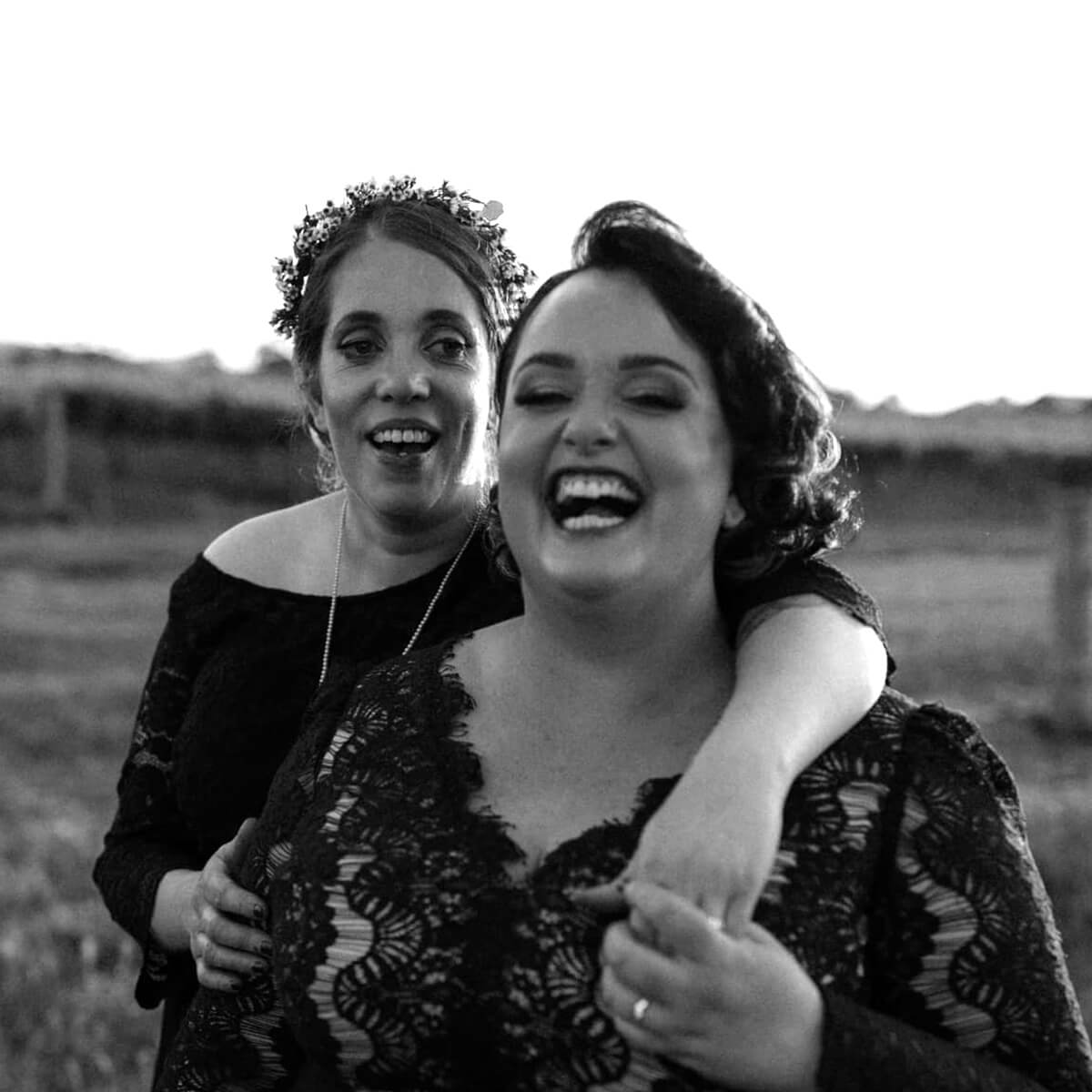 Two women laughing together outdoors, one wearing a floral crown and both in black lace dresses.