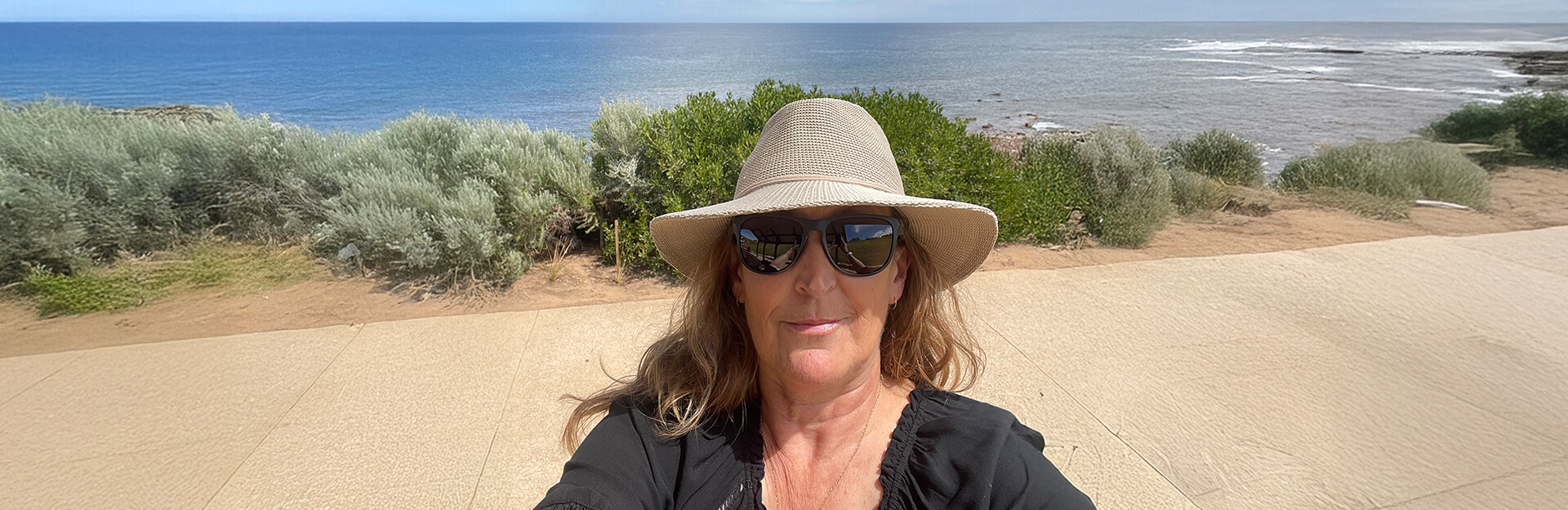 A woman with sunglasses and a wide-brimmed hat poses for a selfie by the ocean, surrounded by greenery.