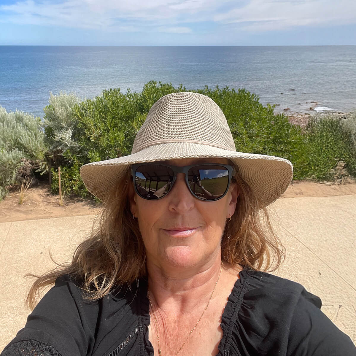 A woman with sunglasses and a wide-brimmed hat poses for a selfie, with the ocean and greenery in the background.