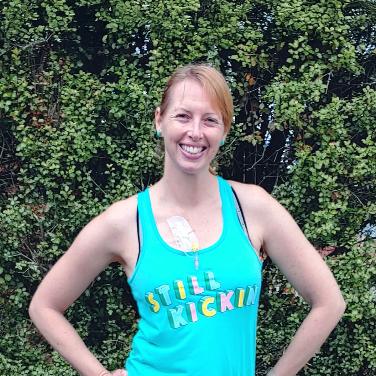 A smiling woman in a turquoise tank top with "STILL KICKIN" printed on it, standing against a green leafy background.