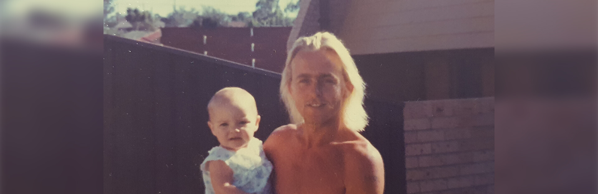 A man holding a baby outdoors, smiling, with a wooden fence and houses in the background.