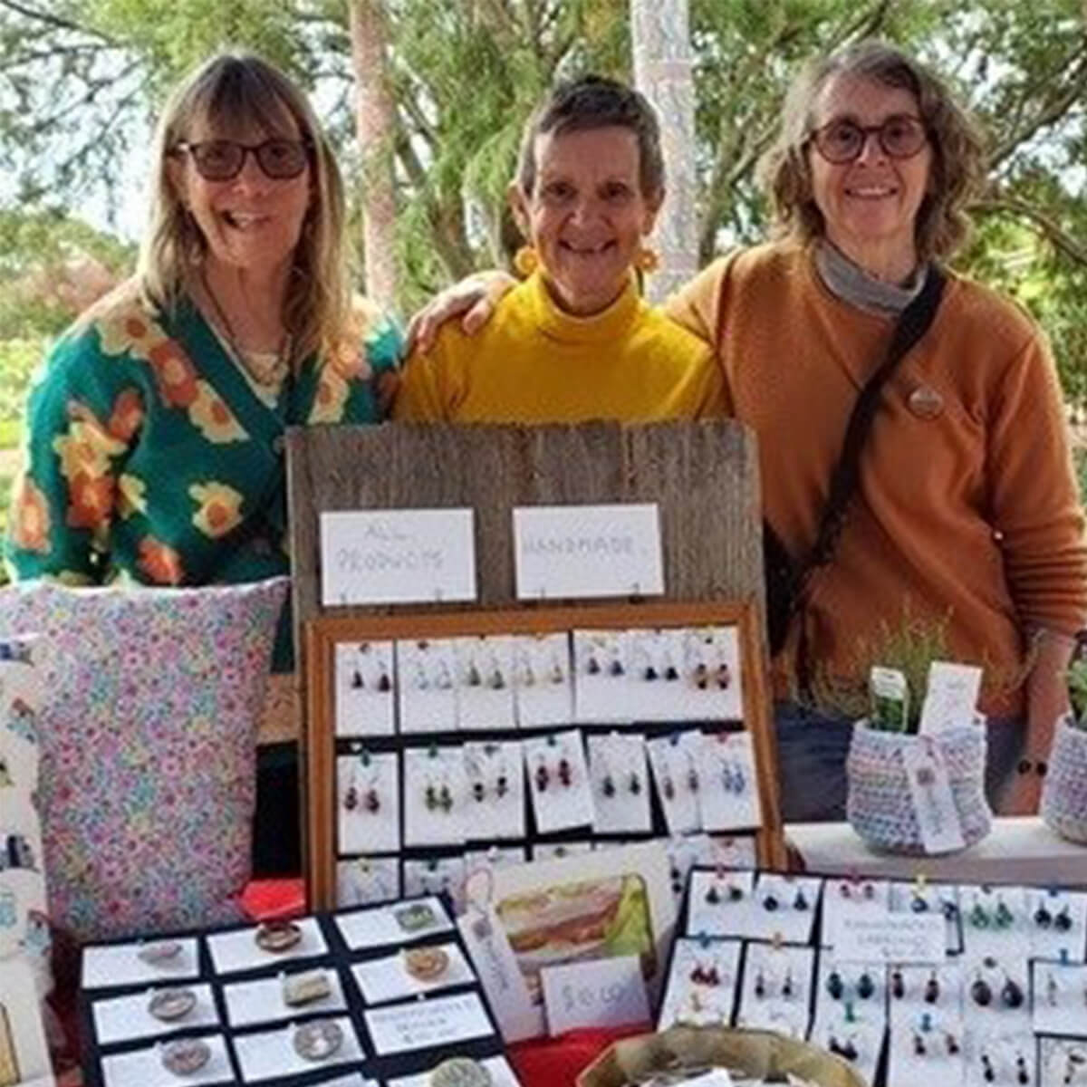 Three women smile behind a table displaying handmade jewelry and crafts at an outdoor market.