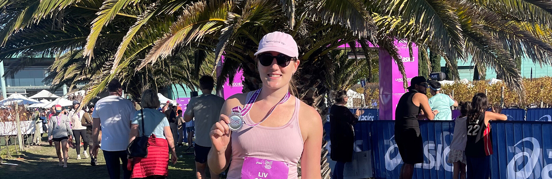 A woman in a pink top and cap holds a medal, surrounded by people and palm trees at an event.