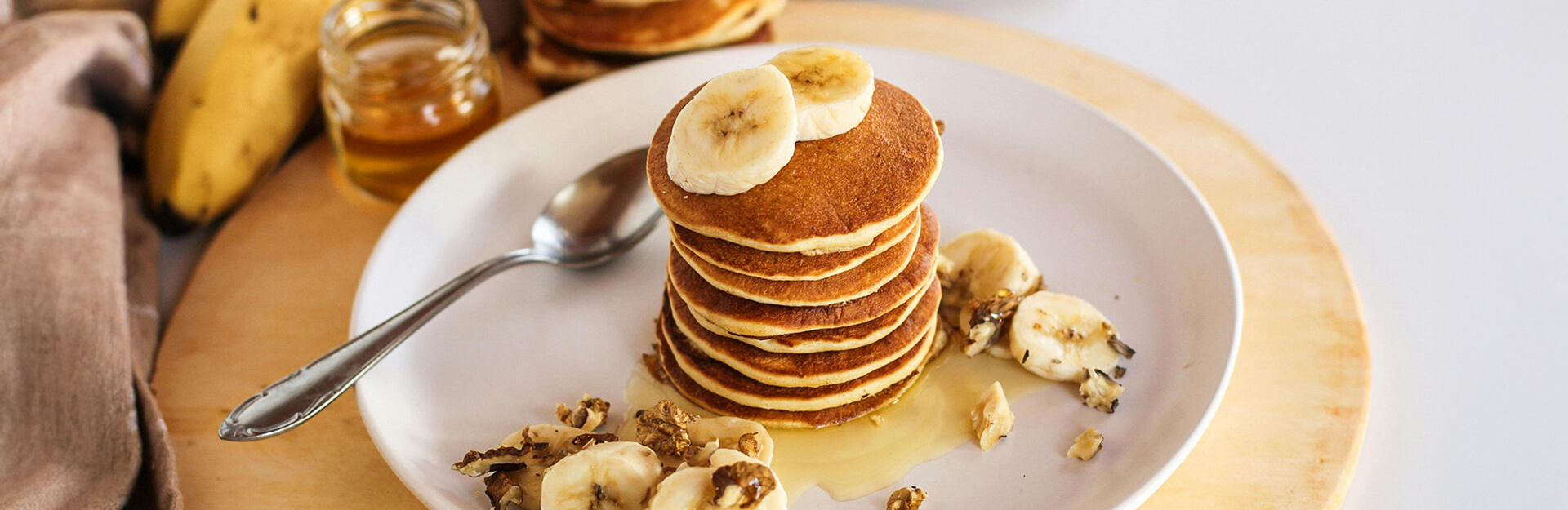A stack of pancakes topped with banana slices and honey, with additional banana pieces and a spoon nearby.