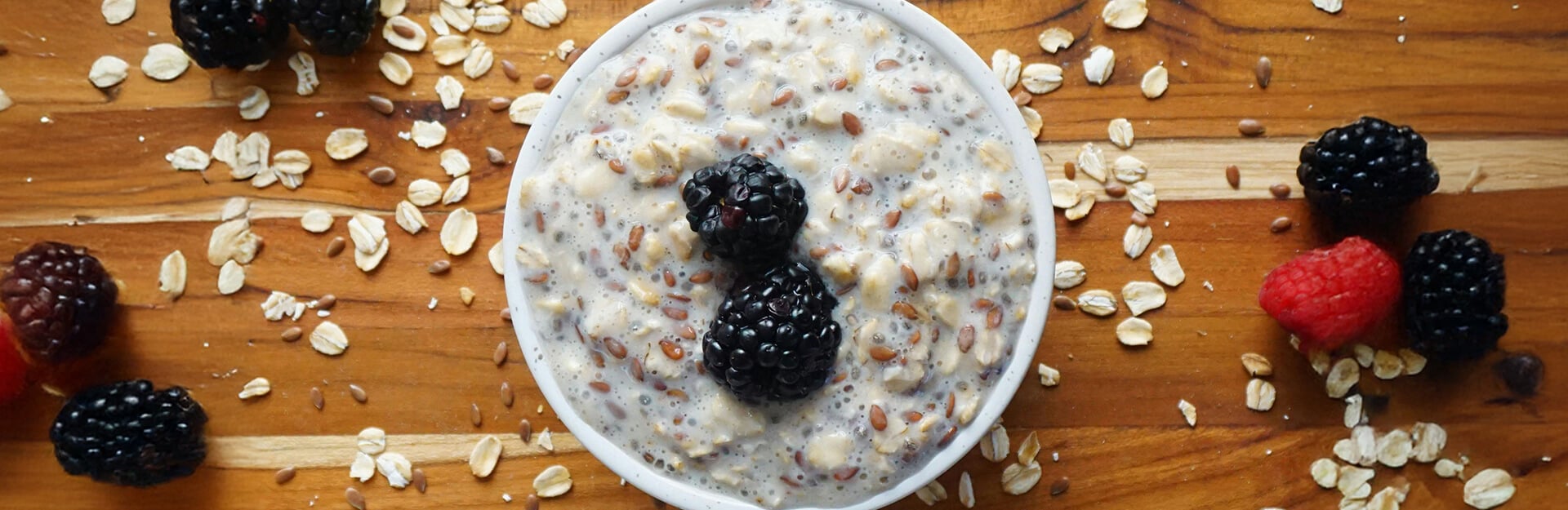 A bowl of oatmeal topped with blackberries and surrounded by scattered oats and berries on a wooden surface.