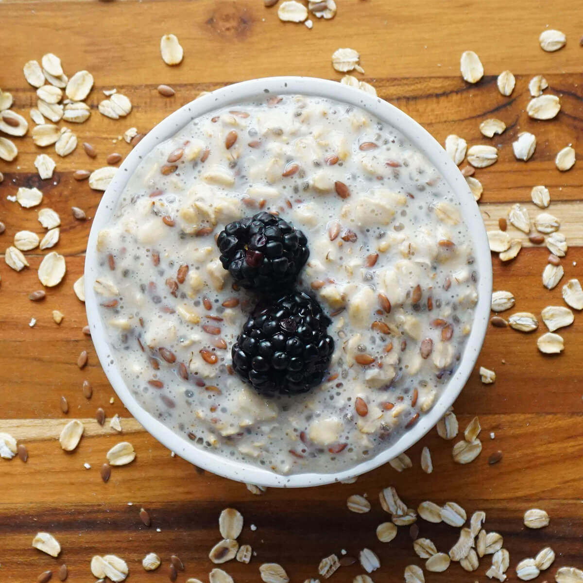 A bowl of oatmeal topped with two blackberries, surrounded by scattered oats and flaxseeds on a wooden surface.