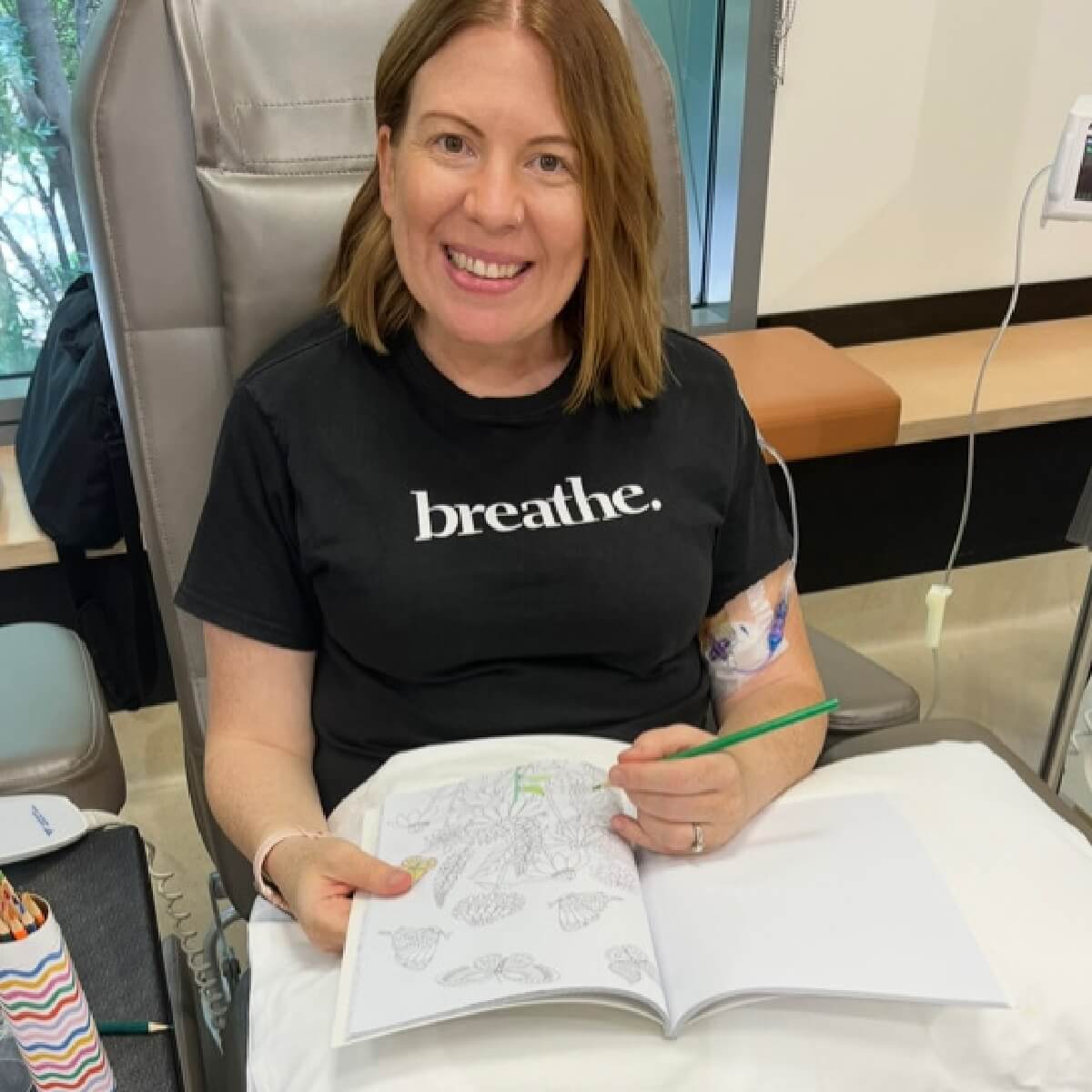 A woman with shoulder-length hair smiles while coloring in a book, wearing a black shirt that says "breathe."