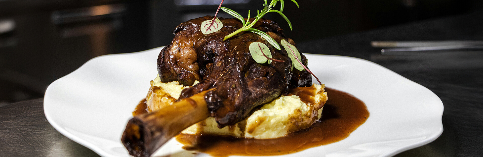 A plated dish featuring a braised lamb shank on mashed potatoes, garnished with microgreens and sauce.