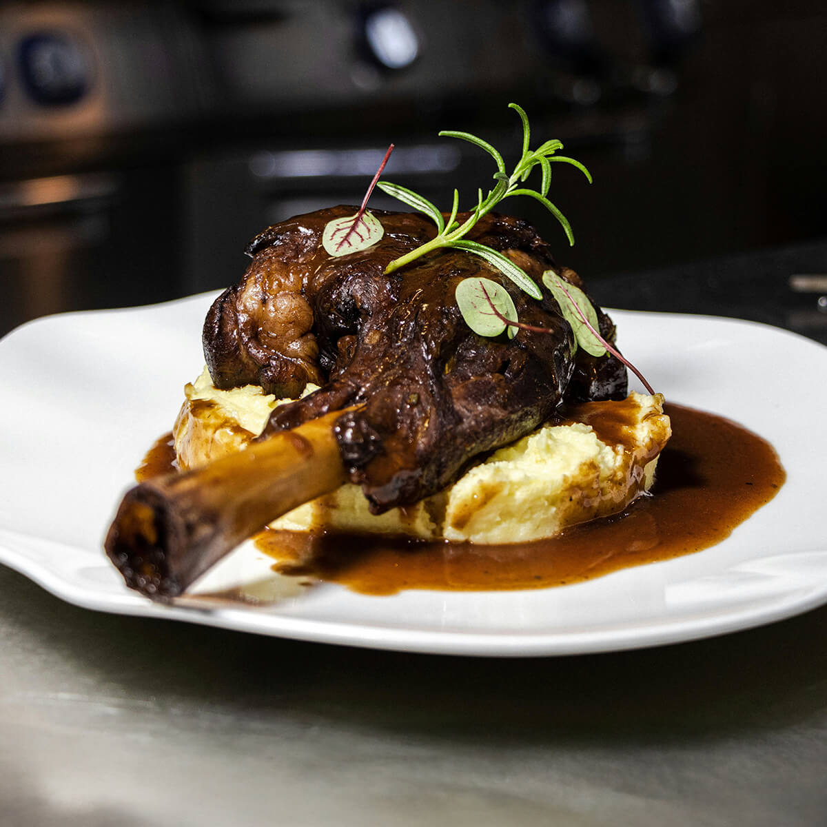 A plated dish featuring a braised lamb shank on creamy mashed potatoes, garnished with microgreens and sauce.