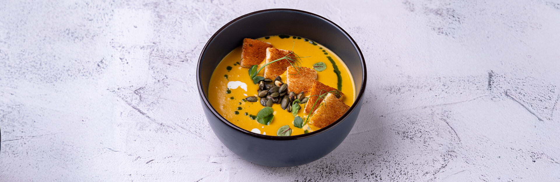 A black bowl filled with creamy orange soup, topped with croutons, seeds, and green herbs on a light textured background.
