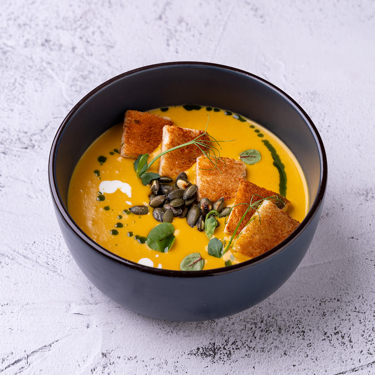 A bowl of creamy orange soup topped with croutons, pumpkin seeds, and green drizzle, garnished with herbs.