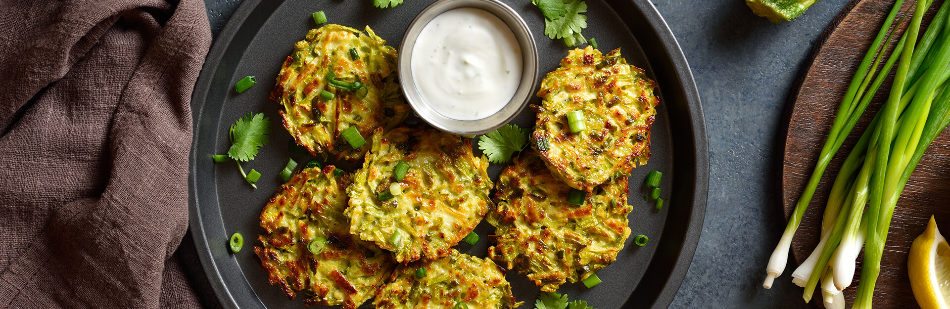 A plate of green veggie fritters with a bowl of dip, garnished with cilantro and green onions, on a textured surface.