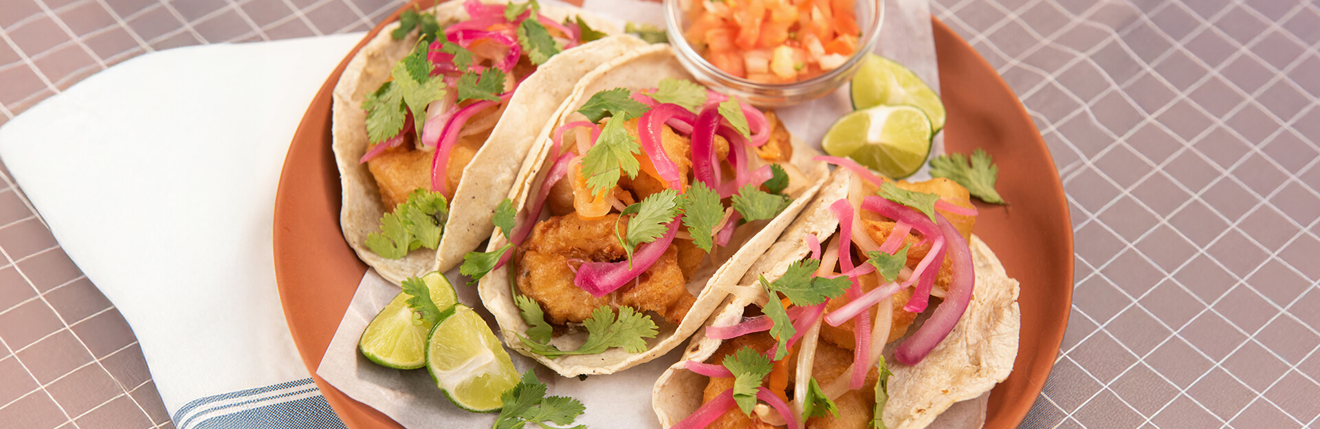 Three tacos filled with fish, topped with cilantro, pickled onions, and served with lime wedges and a bowl of salsa.