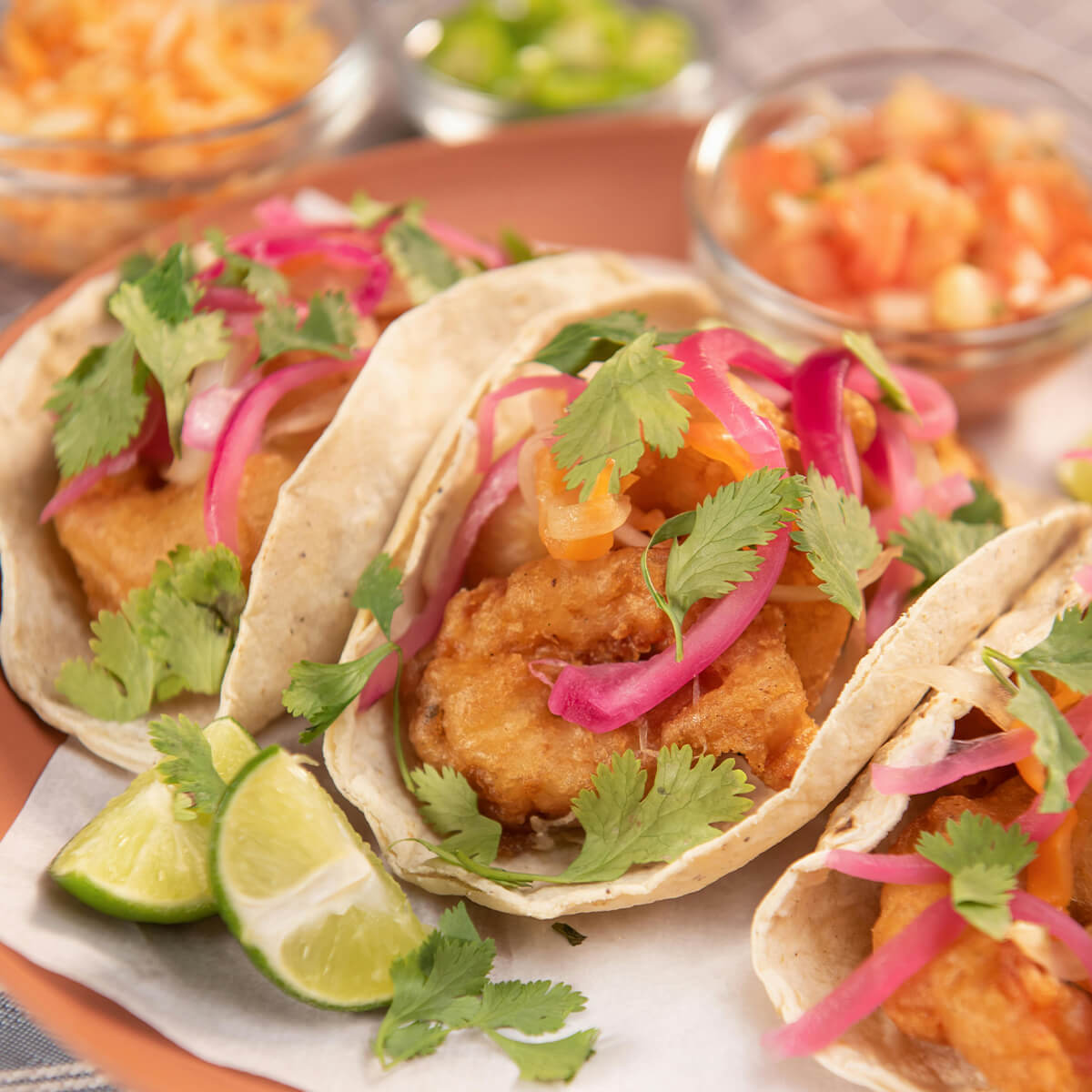 Three tacos filled with fried fish, topped with pink onions and cilantro, served with lime wedges and sides of salsa.