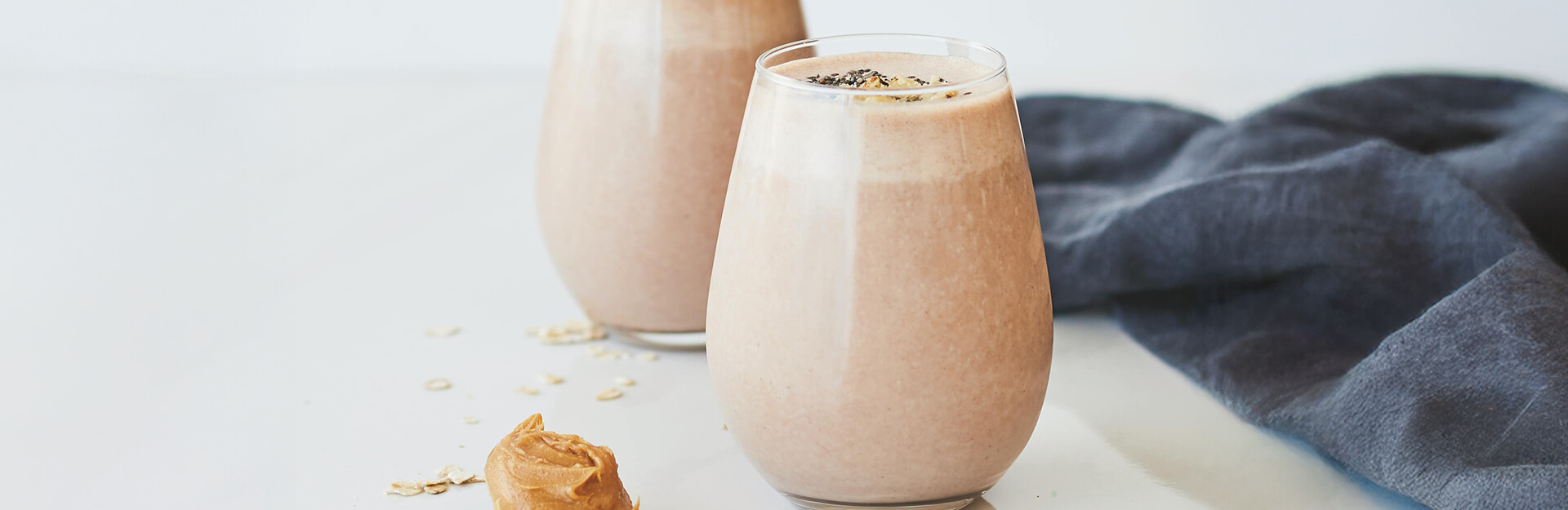 Two glasses of smoothie with a creamy texture, a dollop of peanut butter, and scattered oats on a light surface.