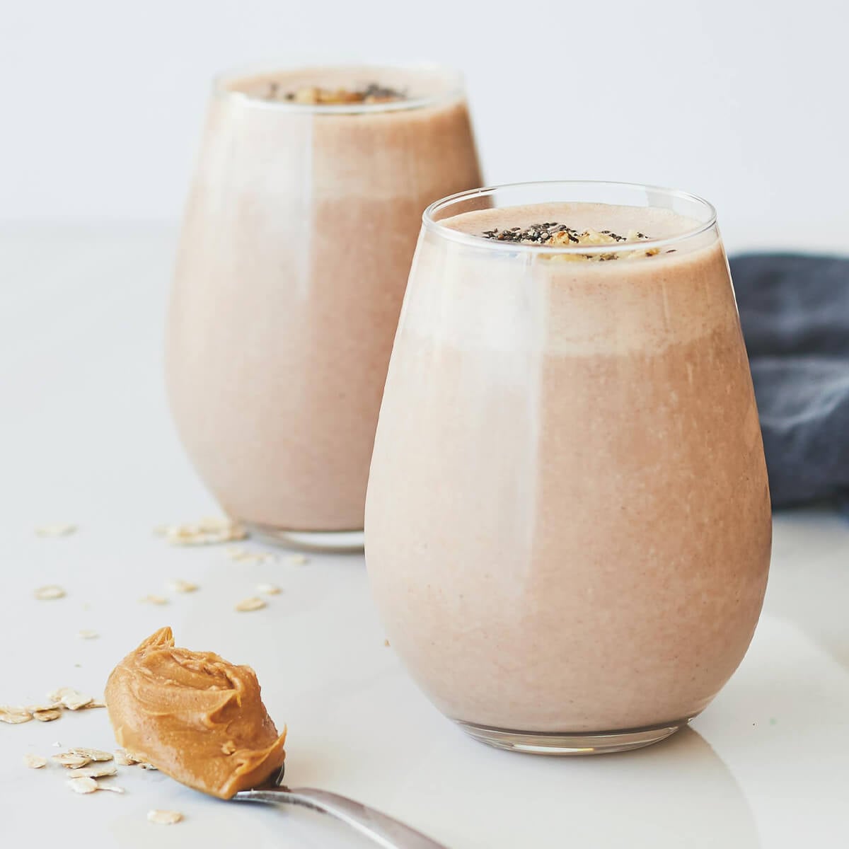 Two smoothies in clear glasses, topped with seeds, beside a spoonful of peanut butter and scattered oats.