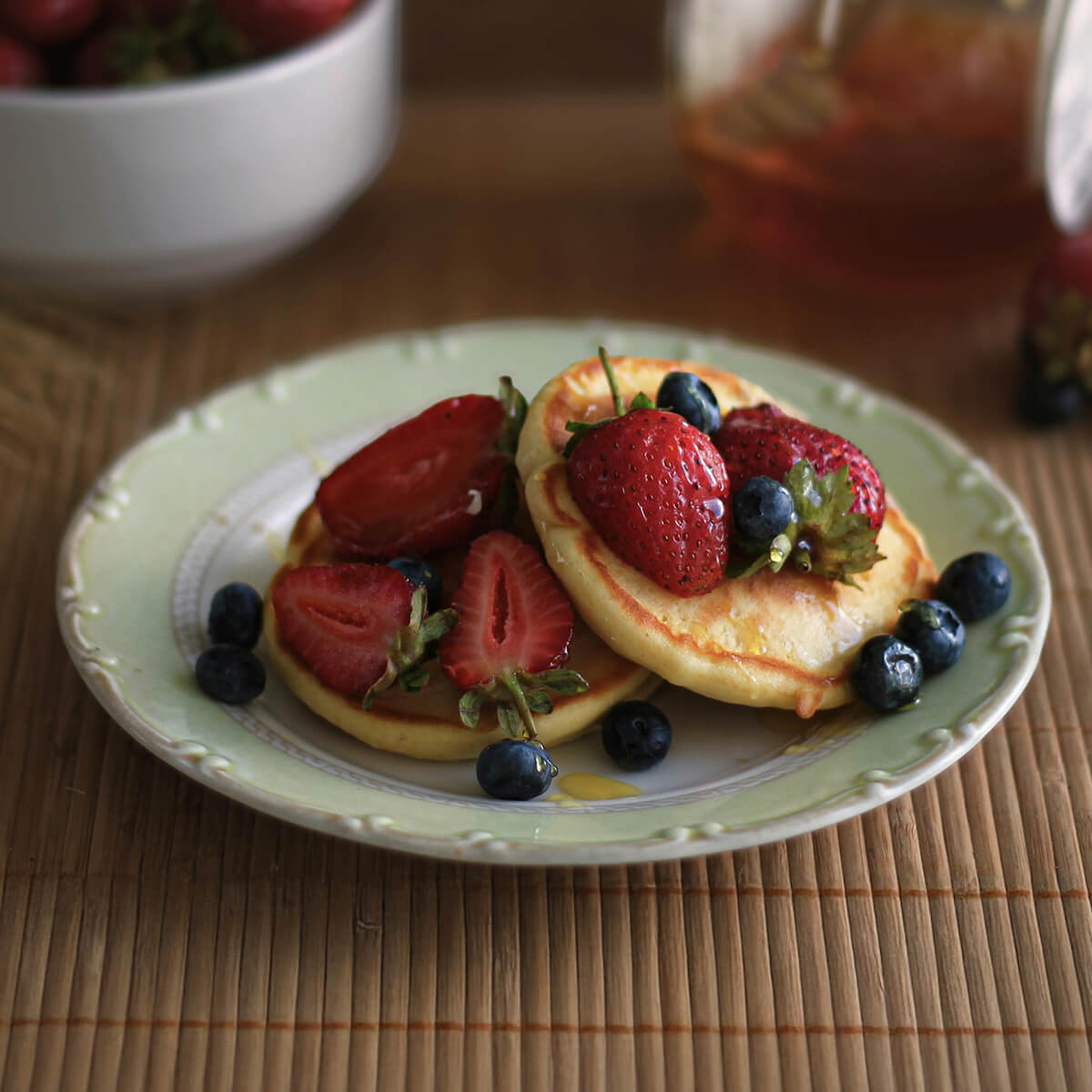 A plate of pancakes topped with strawberries and blueberries, drizzled with syrup, on a bamboo mat.