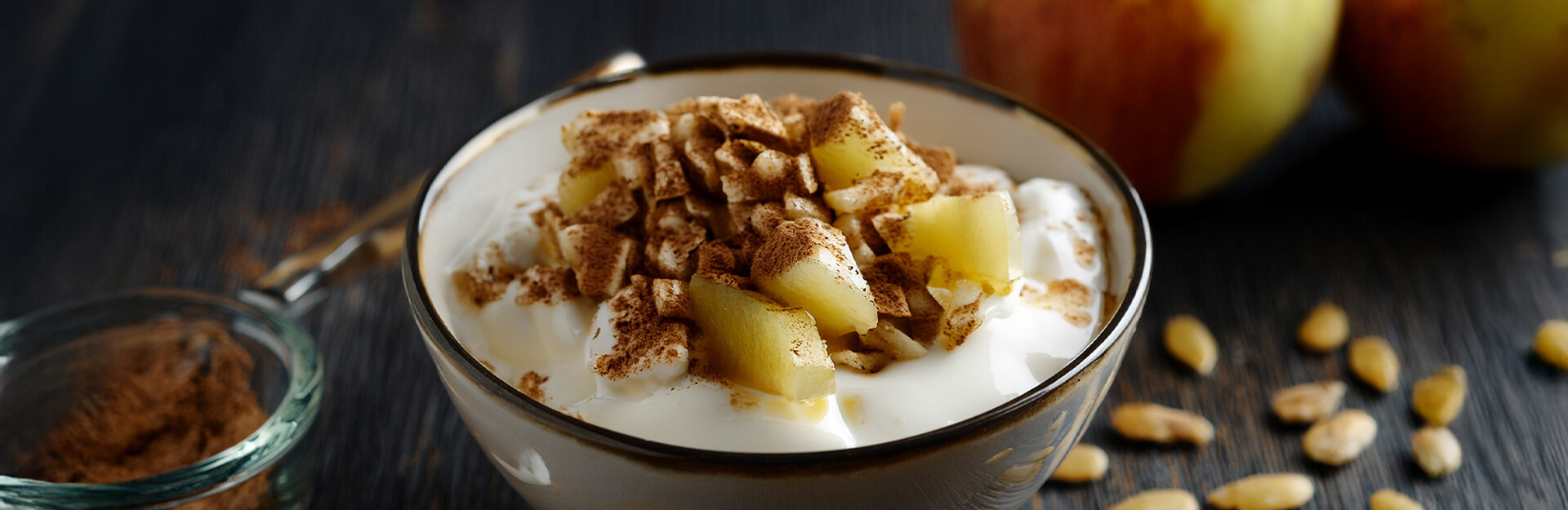 A bowl of yogurt topped with diced apples and cinnamon, with whole apples and seeds in the background.