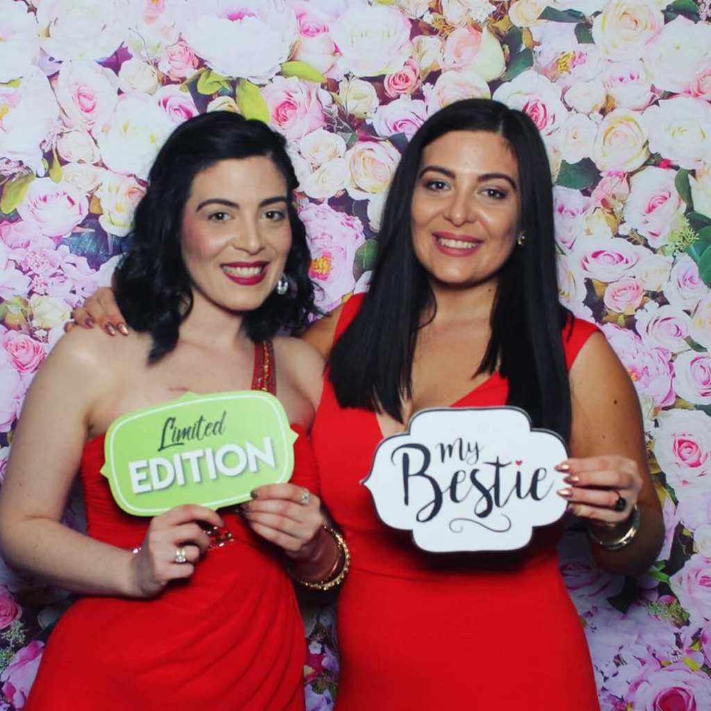 Two women in red dresses pose together, holding signs that read "Limited Edition" and "my Bestie" against a floral backdrop.