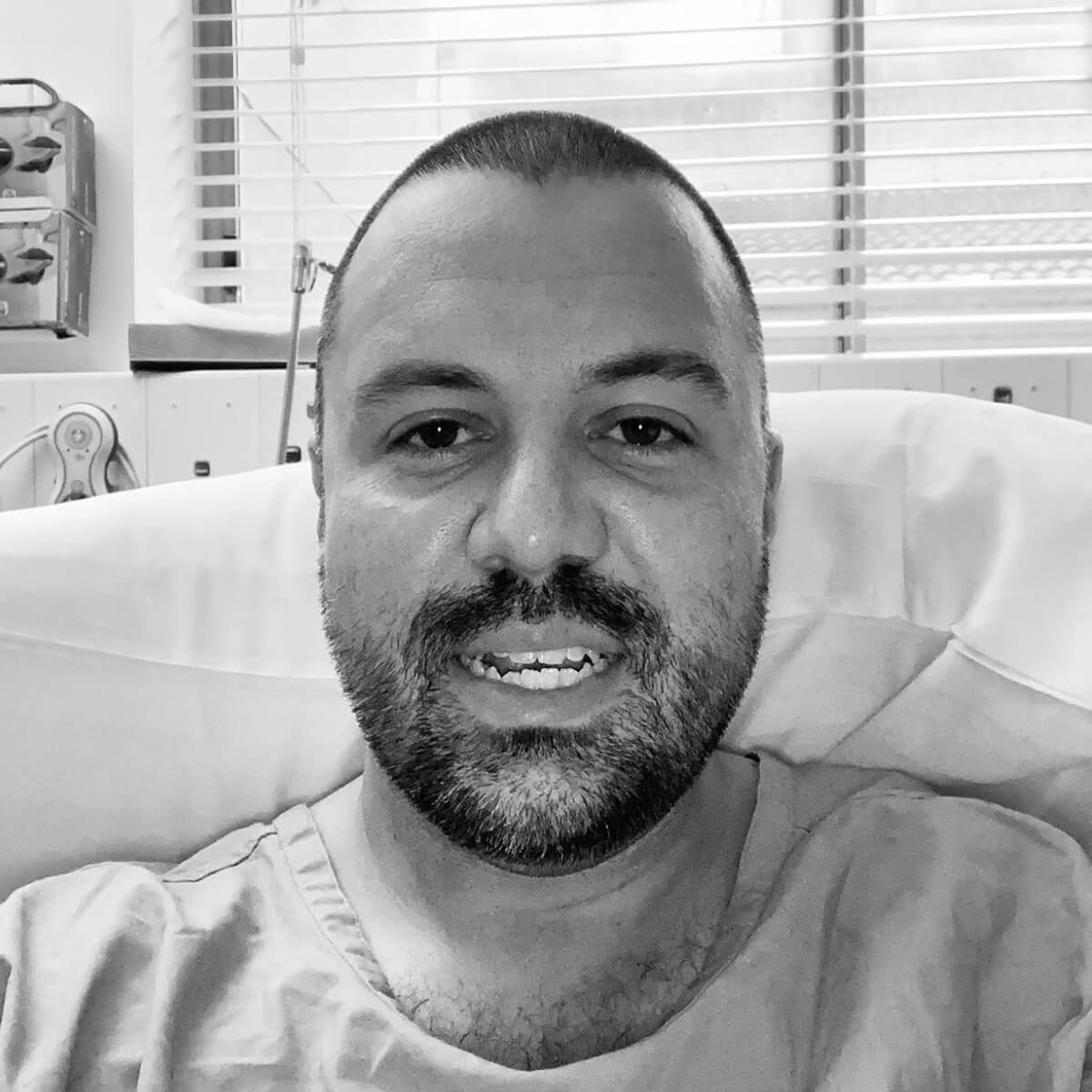 A man in a hospital gown smiles while sitting on a bed, with medical equipment and a window in the background.