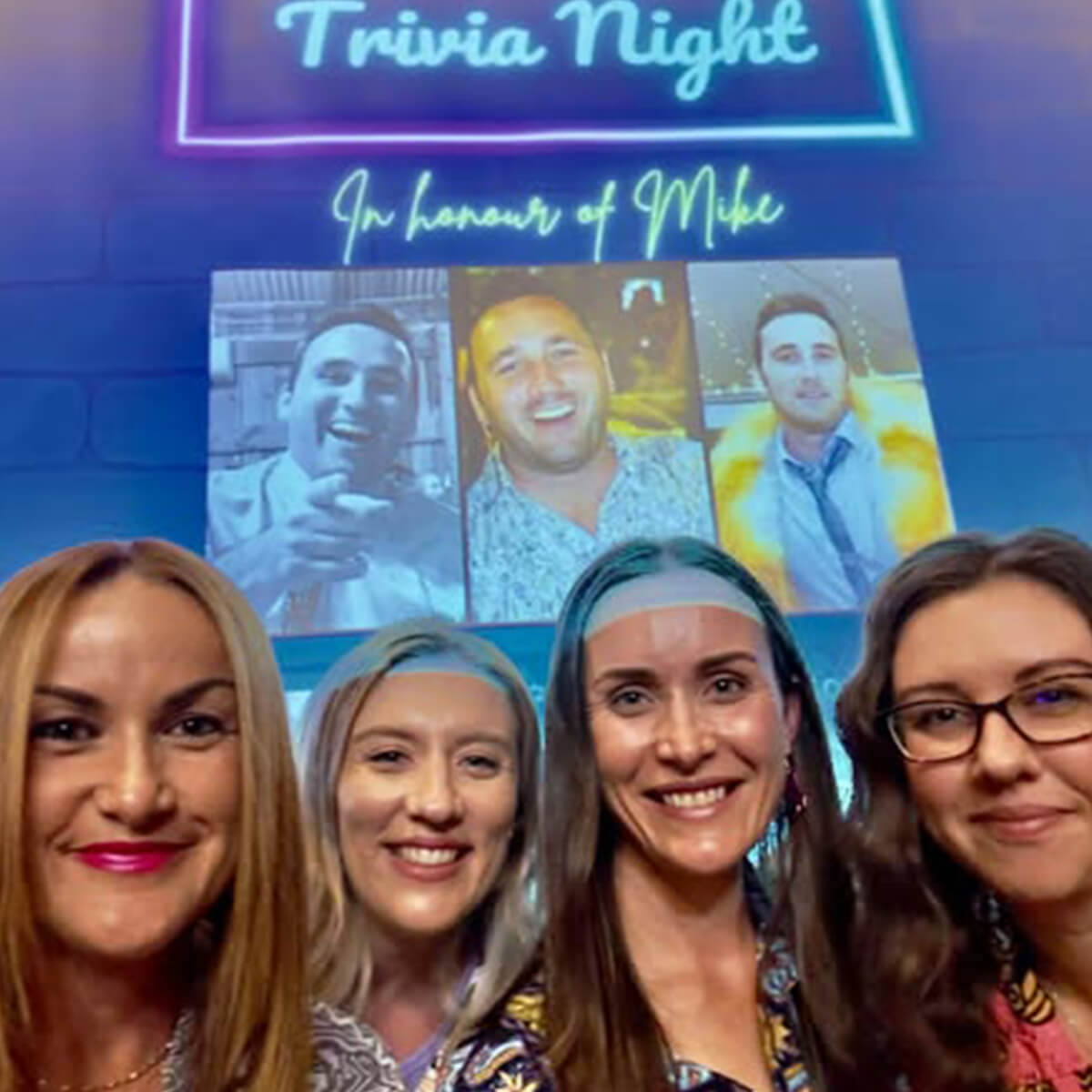 Five women smile for a photo in front of a screen displaying "Trivia Night In honour of Mike" and photos of a man.