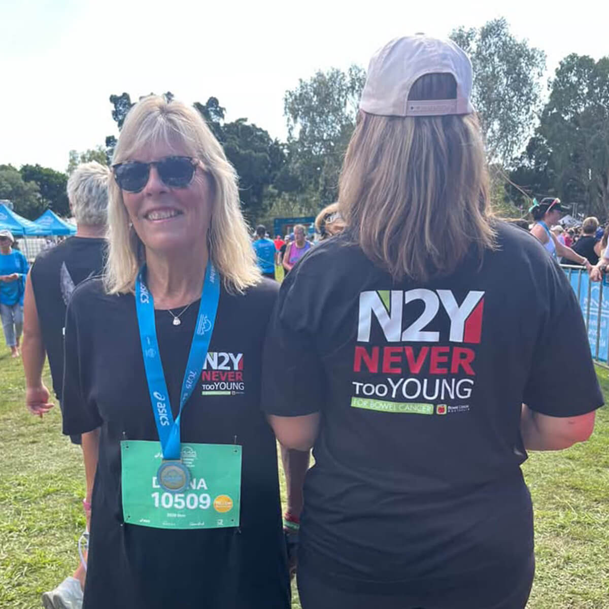 Two women in black t-shirts with "N2Y NEVER tooYOUNG" text, one wearing sunglasses and a medal, at an outdoor event.